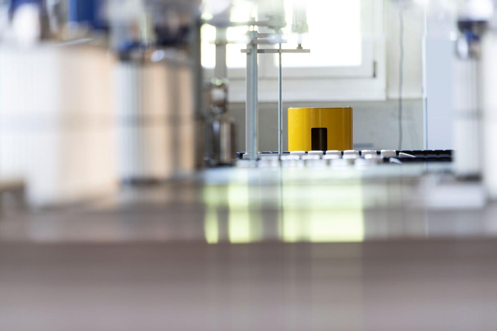 Ein gelbes zylindrisches Objekt befindet sich in der Mitte eines Labors mit unscharfen wissenschaftlichen Geräten und Arbeitsflächen im Vorder- und Hintergrund. Helles Fensterlicht erhellt die Szene.