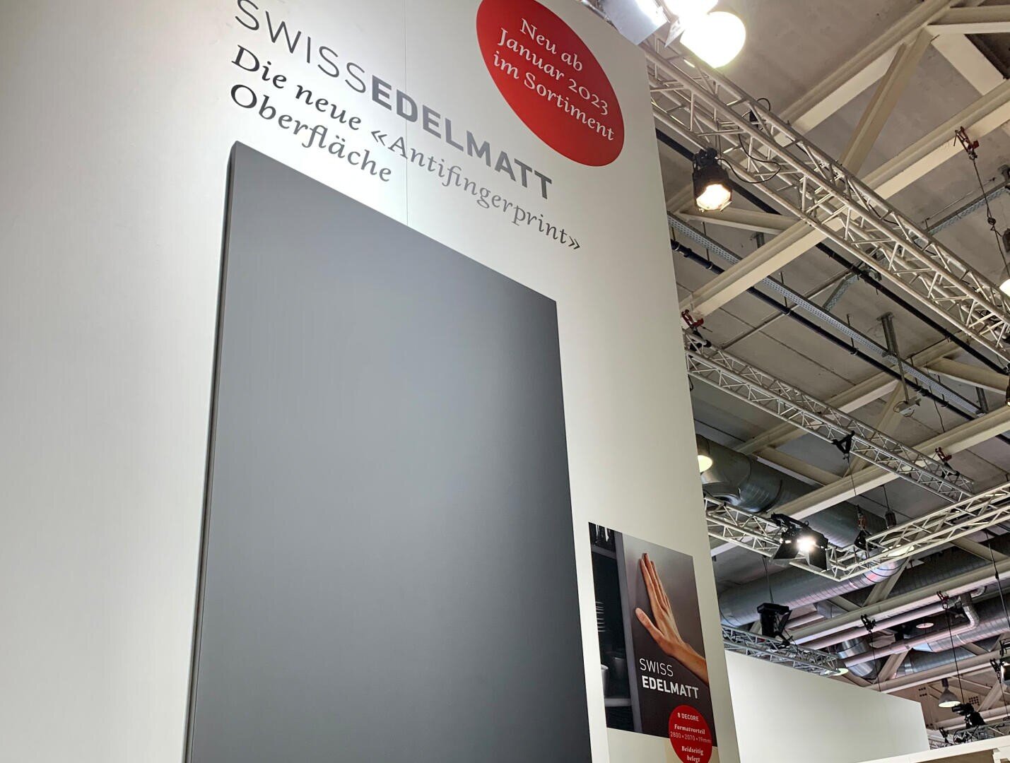 Großes weißes Ausstellungsdisplay mit einer dunklen, matten Platte und deutschem Text, der für eine neue Antifingerprint-Oberfläche ab Januar 2023 von SWISSEDELMATT wirbt. Oben sind Metalltraversen und Deckenleuchten zu sehen.