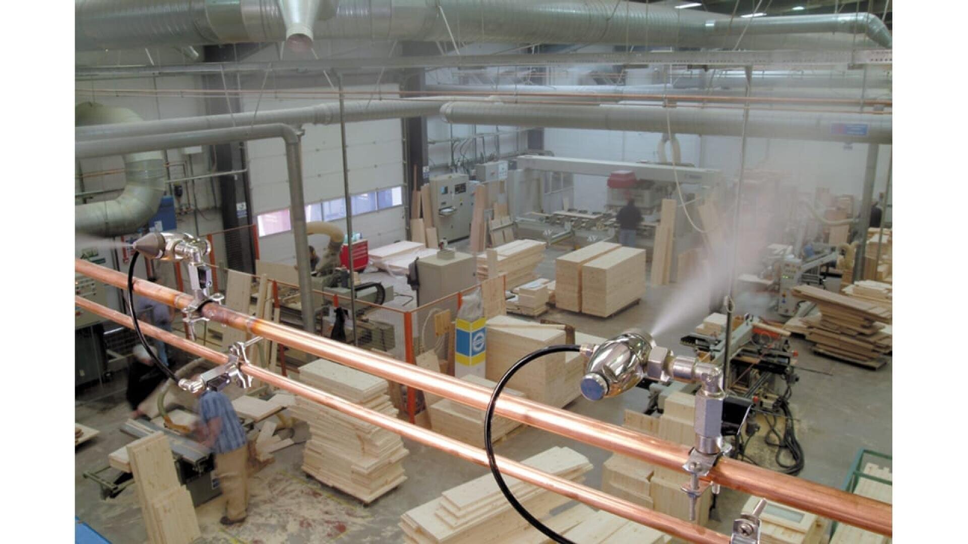 Un grand atelier de menuiserie avec du bois empilé, des établis et des machines, tandis que de la brume s'échappe de tuyaux situés au-dessus de la tête. Plusieurs personnes travaillent parmi les pièces de bois et les équipements dans cet espace industriel lumineux.