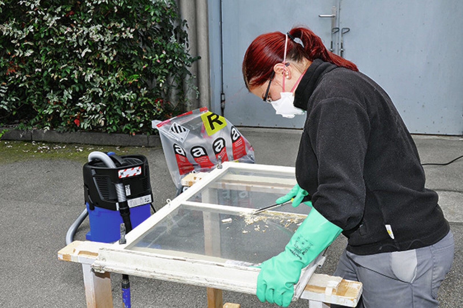 Une personne aux cheveux roux, portant un masque, des gants et une veste noire, gratte la peinture d'un cadre de fenêtre sur un établi à l'extérieur. Il y a des sacs de matériaux et un aspirateur d'atelier à proximité.