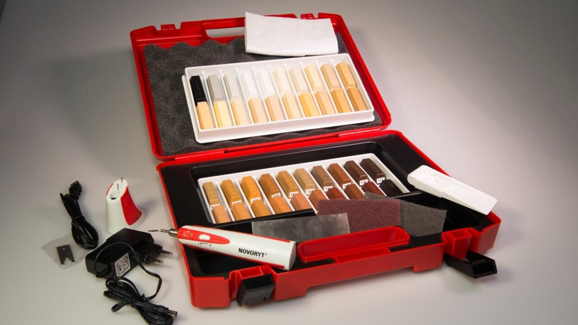 Une mallette en plastique rouge contenant des bâtons de mastic à bois de différentes teintes, du papier de verre, un chiffon blanc, un outil chauffant électrique, un chargeur et d'autres accessoires de réparation disposés sur une surface grise.