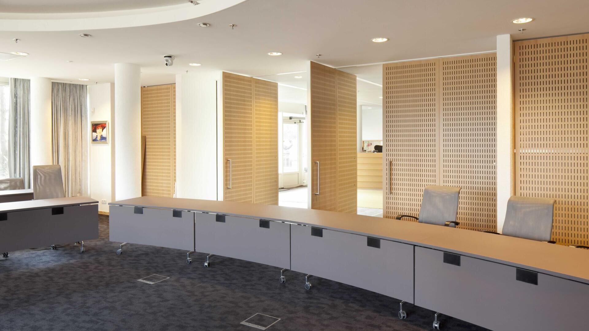 Moderner Konferenzraum mit einem langen, geschwungenen Tisch, mehreren Bürostühlen auf Rollen, hellen Holzwänden und großen Fenstern, die natürliches Licht in den Raum lassen.