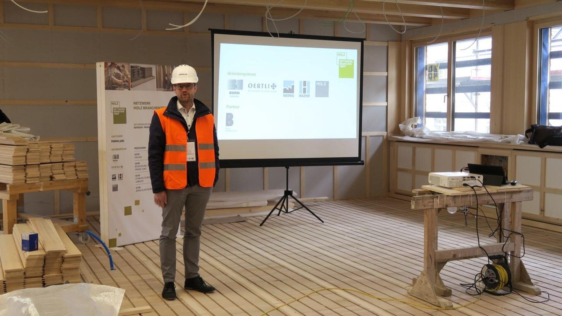 Un homme portant un casque blanc, des lunettes et un gilet de sécurité orange se tient sur un chantier de construction en bois, à côté d'un écran de projecteur affichant des logos d'entreprise. Des piles de planches de bois et de matériaux de construction sont visibles.