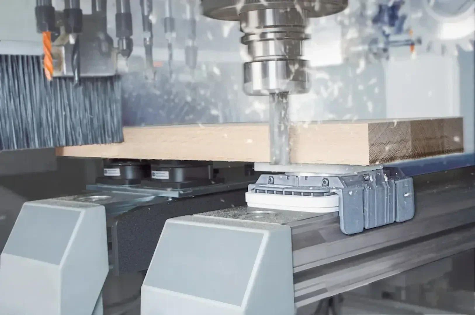 Eine CNC-Maschine schneidet ein rechteckiges Holzstück, wobei Metallklammern das Holz in Position halten und Sägespäne aufgewirbelt werden, während die Bohrmaschine arbeitet. Oberhalb des Holzes sind verschiedene Schneid- und Bohrwerkzeuge zu sehen.