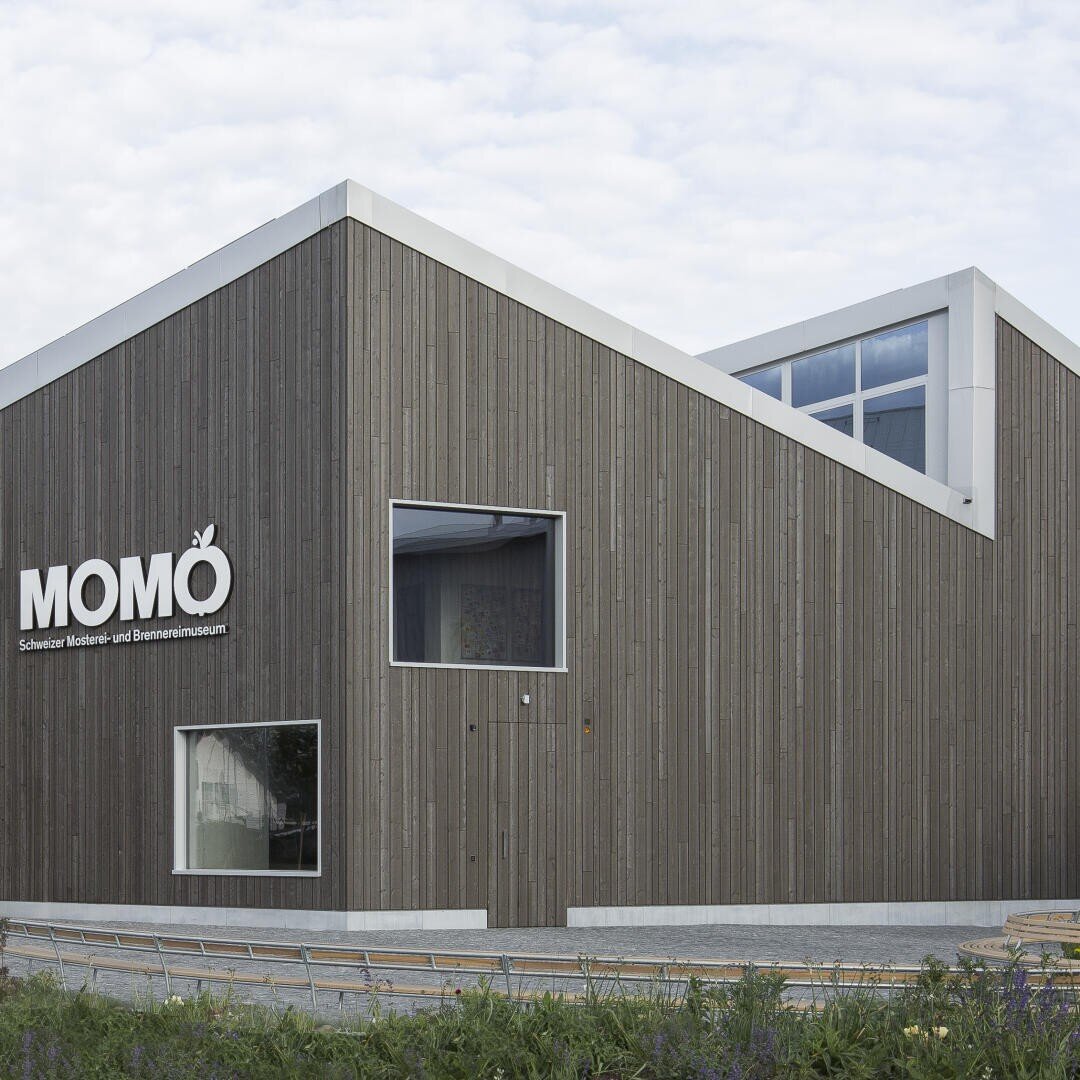 Ein modernes Holzgebäude mit kantigen Dächern hat große Fenster und ein Schild mit der Aufschrift MOMÖ Schweizer Mosterei- und Brennereimuseum. Im Vordergrund befinden sich Grünflächen und ein geschwungener Weg.