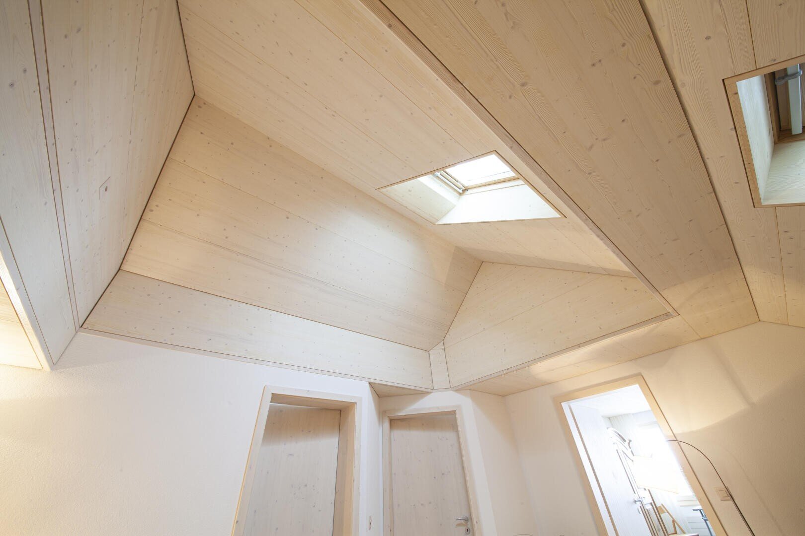 Eckansicht eines modernen Raums mit Deckenpaneelen aus hellem Holz, die geometrische Formen aufweisen, und einem Oberlicht, das über zwei geschlossenen Türen natürliches Licht hereinlässt.