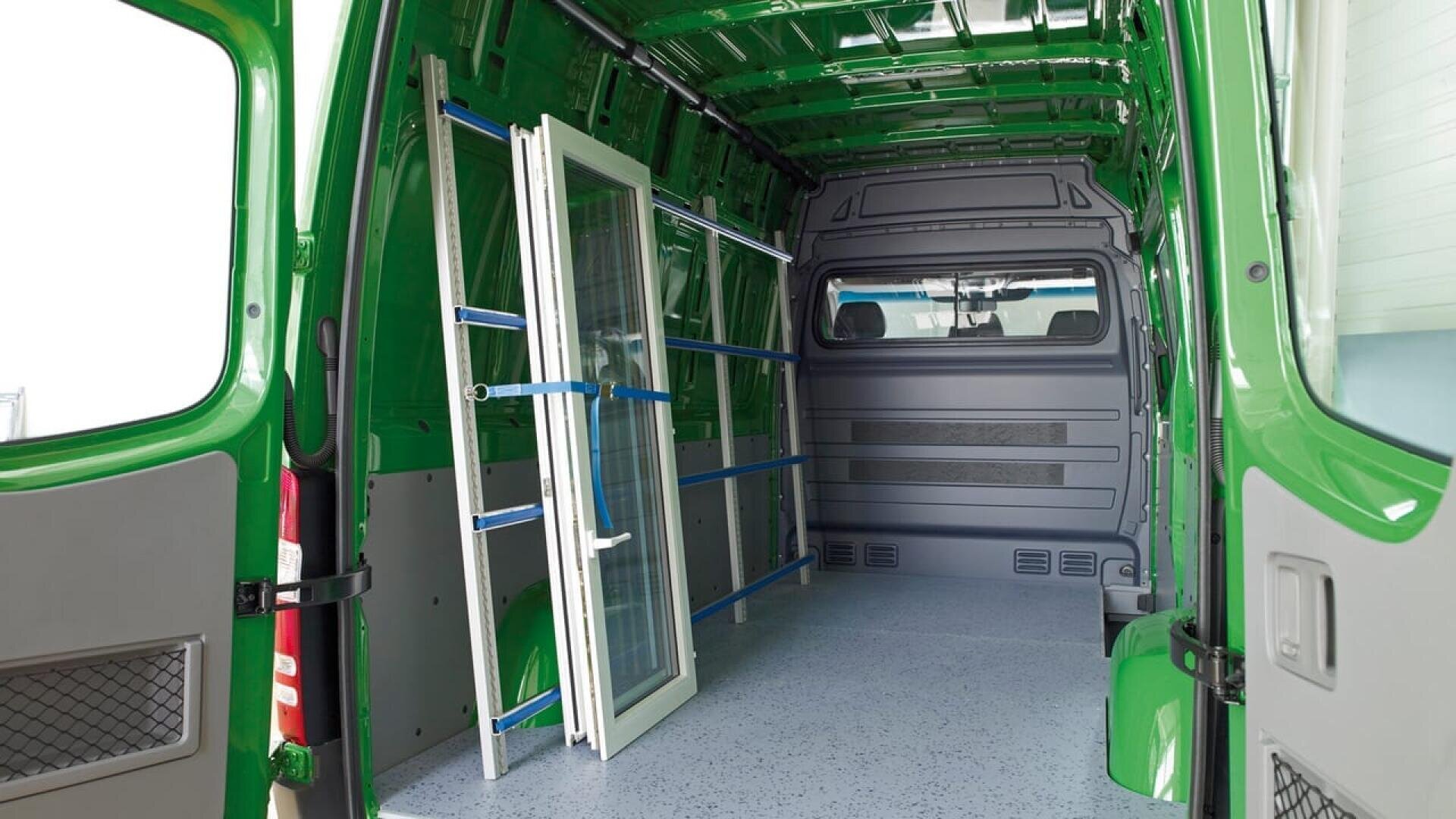 Intérieur d'une camionnette de livraison verte dont l'espace de chargement est vide, à l'exception d'une grande fenêtre ou d'un cadre de porte en verre fixé dans un support métallique bleu le long de la paroi latérale gauche. La camionnette semble propre et bien organisée.