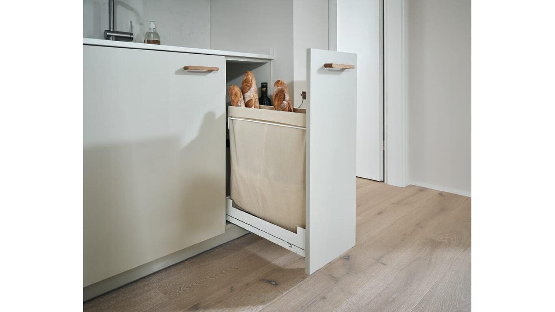 Un meuble de cuisine avec une corbeille en tissu coulissante contenant plusieurs baguettes ; le meuble est de couleur claire avec des poignées en bois et repose sur un sol en bois clair.