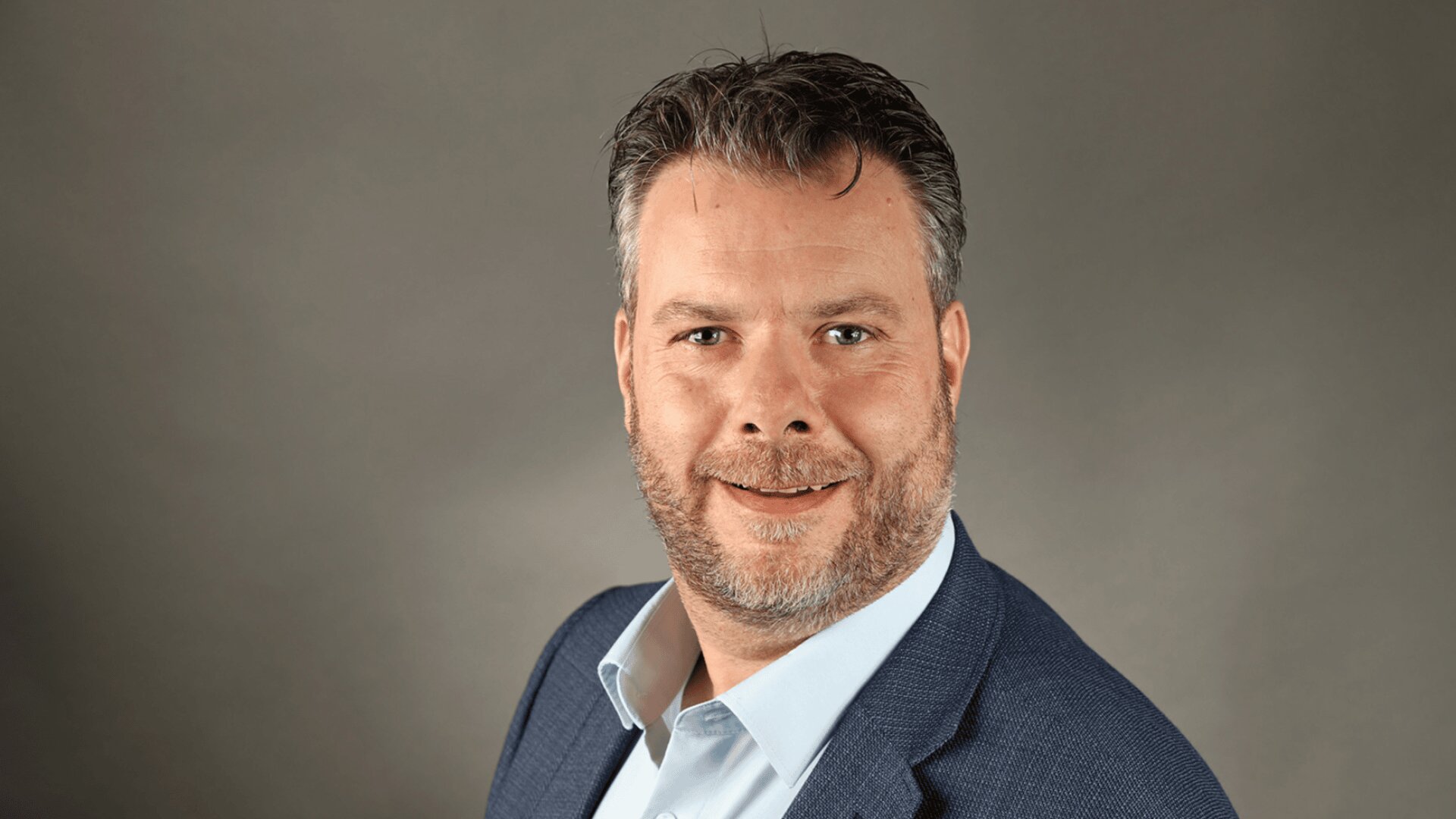 Ein Mann mittleren Alters mit kurzen braunen Haaren, Bart und Schnurrbart, der einen blauen Blazer und ein hellblaues Hemd trägt, lächelt vor einem einfarbig grauen Hintergrund in die Kamera.