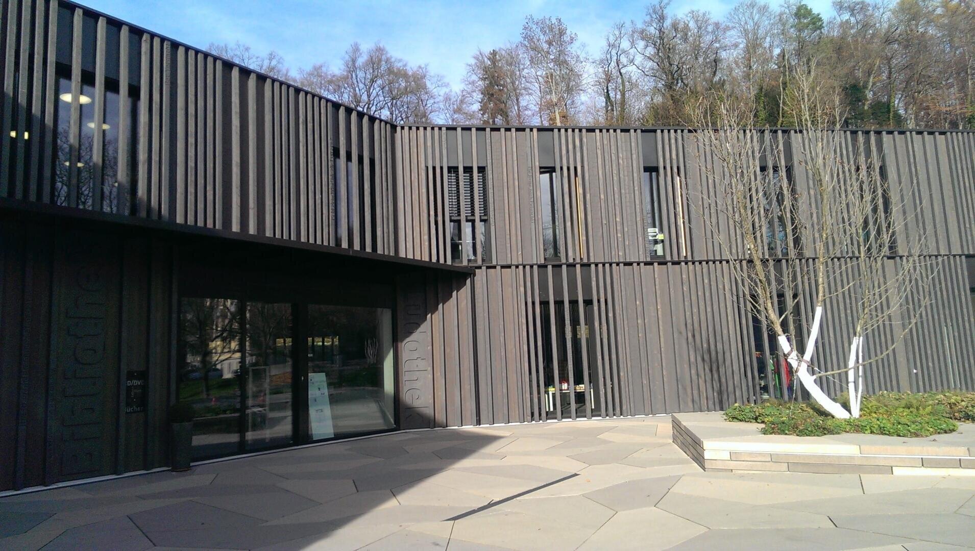 Ein modernes zweistöckiges Bibliotheksgebäude mit vertikalen Holzlatten an der Außenseite, großen gläsernen Eingangstüren, geometrischem Pflaster und blattlosen Bäumen an der Vorderseite in der Nähe eines Waldgebiets.