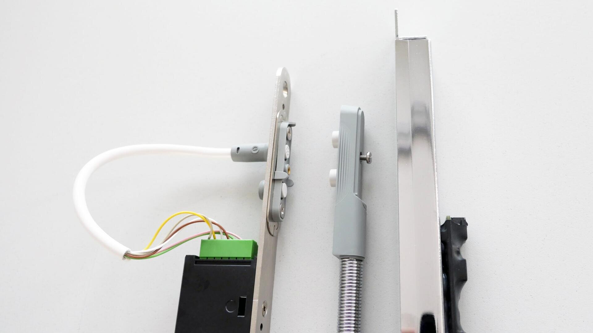 Gros plan sur des composants électroniques et mécaniques d'une serrure, y compris un circuit imprimé avec des fils, un mécanisme de verrouillage en métal et une tige métallique brillante, le tout placé sur une surface gris clair.