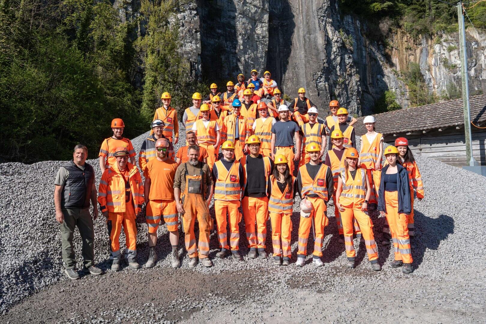 Eine große Gruppe von Personen mit orangefarbenen Sicherheitswesten, Schutzhelmen und Arbeitsausrüstung posiert gemeinsam auf Kies vor einer felsigen Klippe mit Bäumen und einem Holzgebäude im Hintergrund.