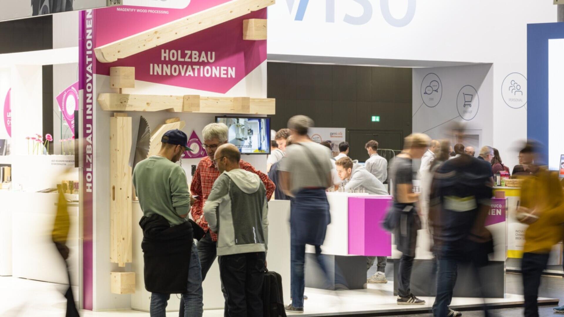 Les gens se rassemblent sur un stand d'exposition intitulé Holzbau Innovationen, qui présente des constructions en bois et des touches de rose vif, tandis que d'autres se promènent dans un espace événementiel intérieur très fréquenté.