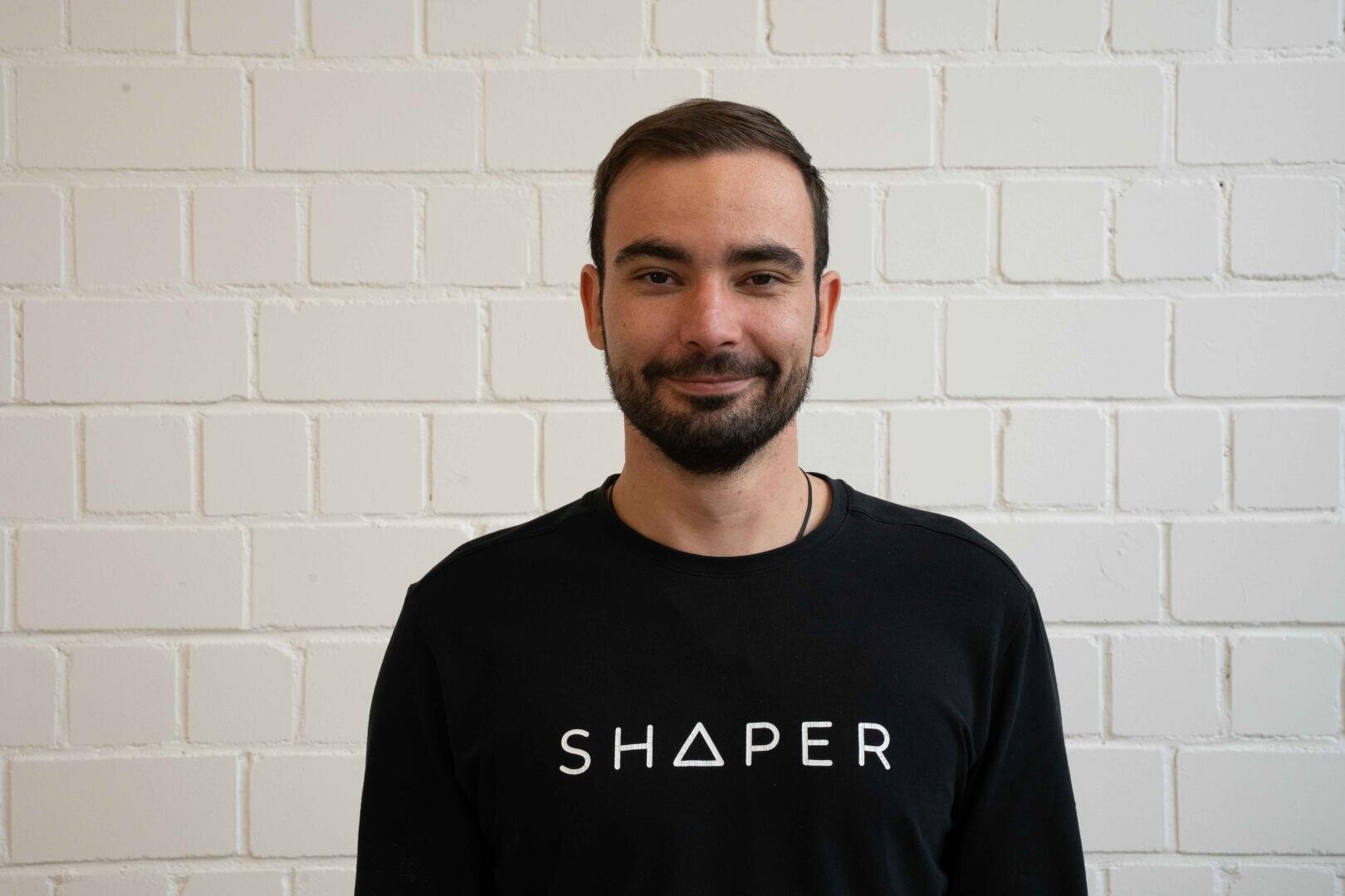 Un homme aux cheveux bruns courts et à la barbe se tient devant un mur de briques blanches et porte une chemise noire sur laquelle est inscrit le mot SHAPER en lettres blanches.