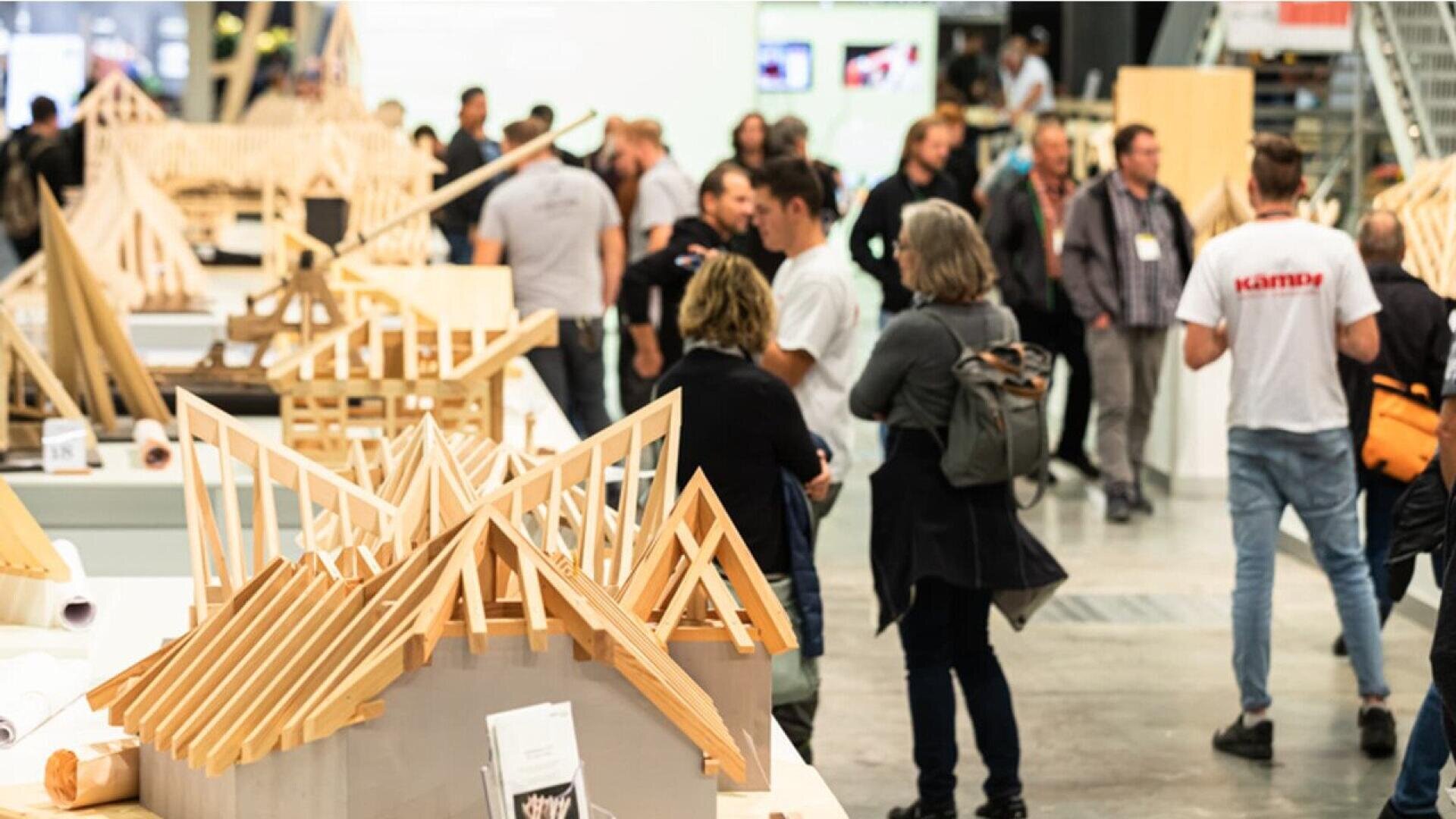 Un groupe de personnes se promène et se rassemble autour de tables présentant des modèles architecturaux en bois lors d'une exposition ou d'un événement en intérieur.