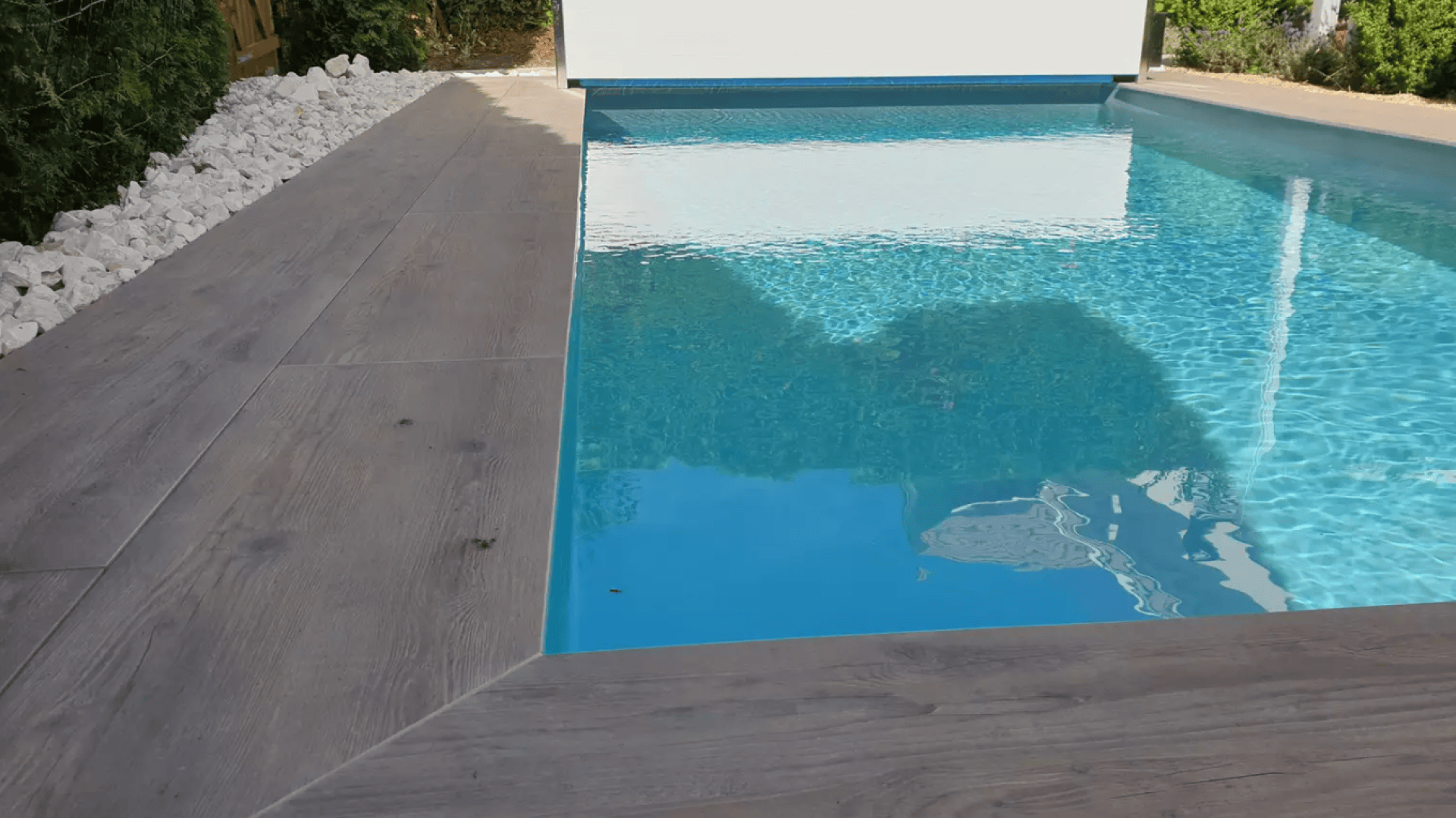 Ein moderner rechteckiger Pool mit hellblauem Wasser, umgeben von einer Terrasse aus hellem Holz und weißen Ziersteinen entlang des Randes, mit Grün im Hintergrund.