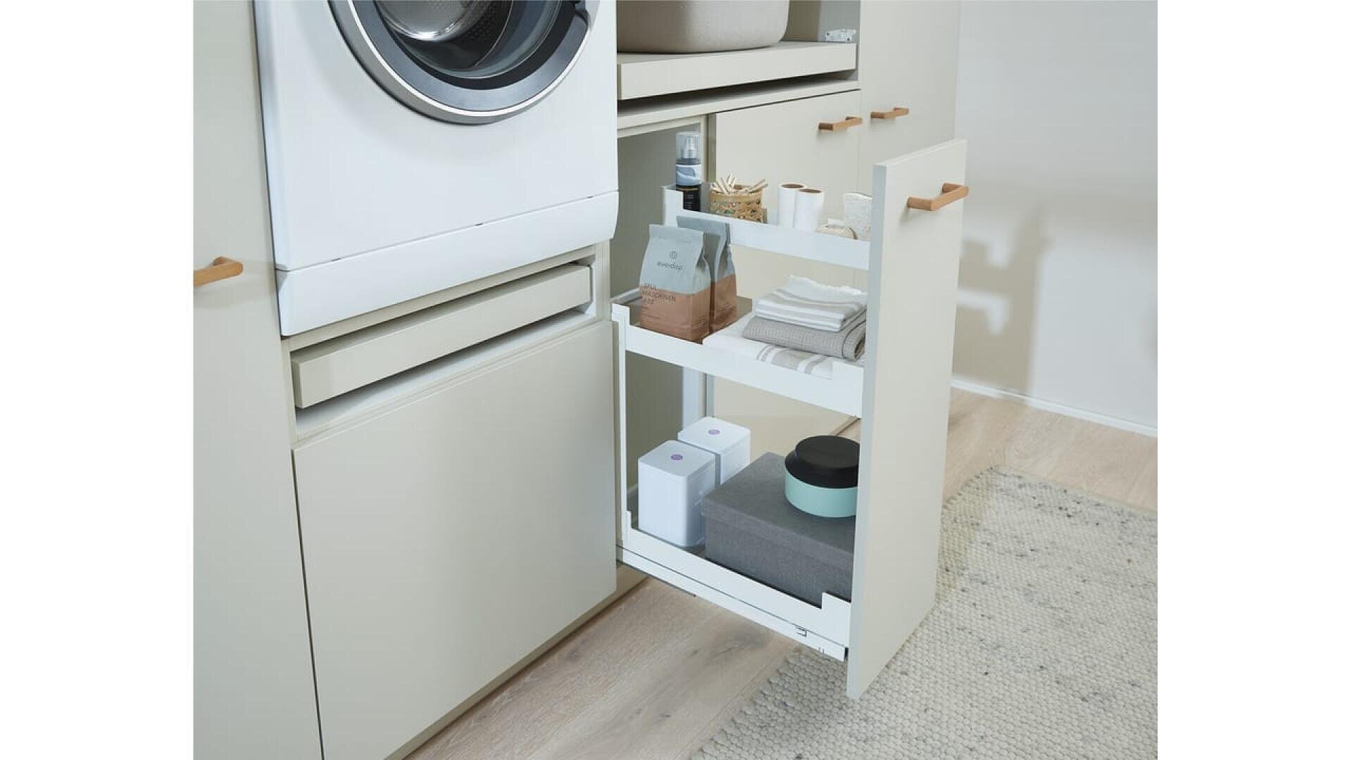 L'espace buanderie est équipé d'un lave-linge à chargement frontal surmonté d'une étagère coulissante contenant des serviettes, des boîtes, des bouteilles et d'autres petites fournitures de blanchisserie. Le sol est en bois clair avec un tapis tissé.