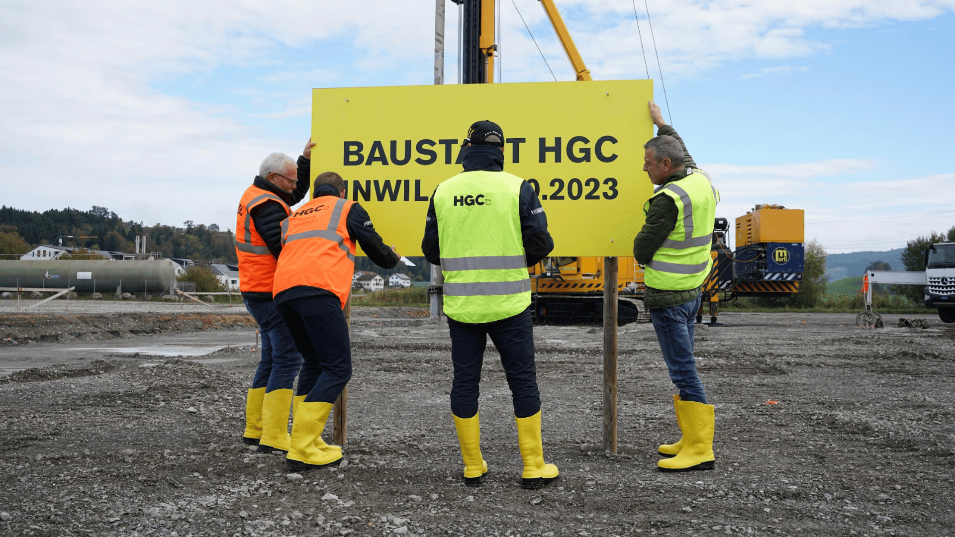 Vier Arbeiter in gelben Stiefeln und Sicherheitswesten stellen ein großes gelbes Schild auf einer Baustelle auf. Auf dem Schild steht BAUSTART HGC INWIL 2023. Im Hintergrund sind Maschinen und ein bewölkter Himmel zu sehen.