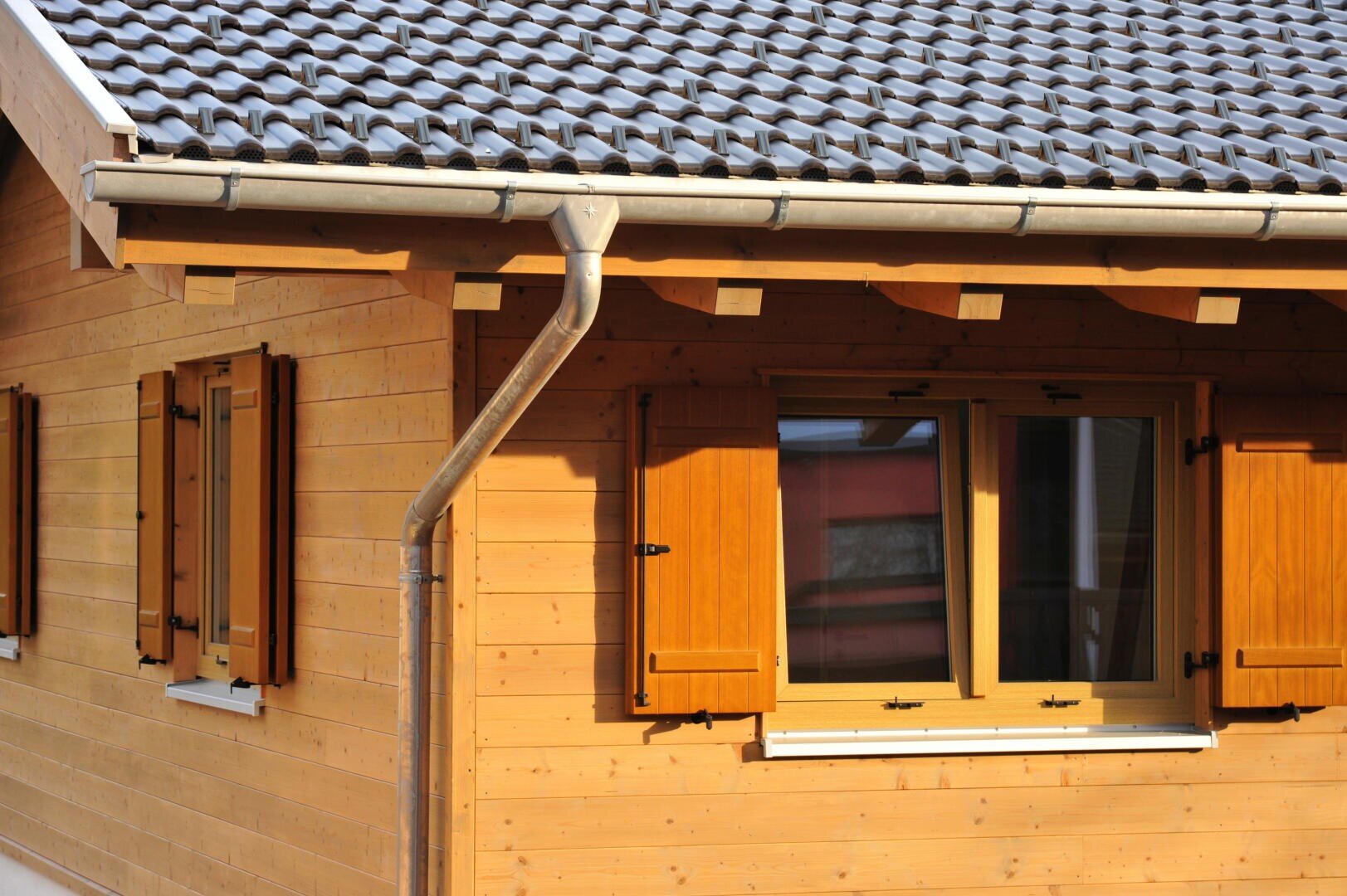 Eine Nahaufnahme eines Holzhauses mit zwei verschlossenen Fenstern, einem Ziegeldach und einer Regenrinne aus Metall an einem sonnigen Tag.