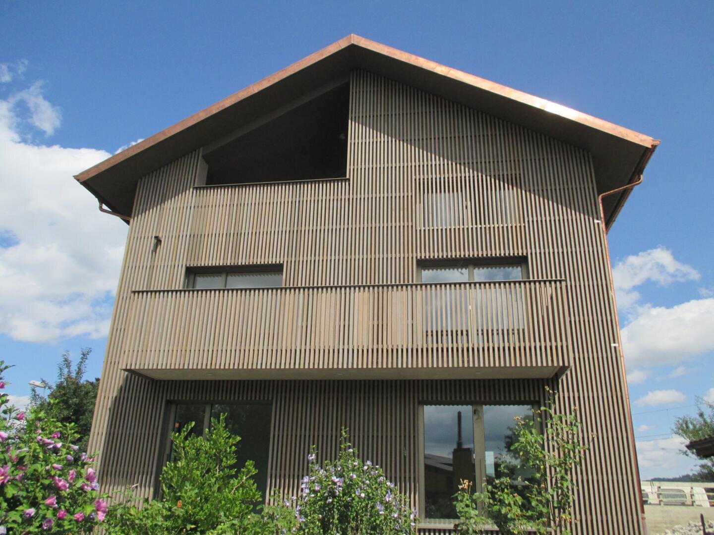 Ein modernes zweistöckiges Haus mit vertikalen Holzlatten an der Fassade, großen Fenstern und einem dreieckigen Dach, umgeben von Grün und blühenden Pflanzen unter einem blauen Himmel mit Wolken.