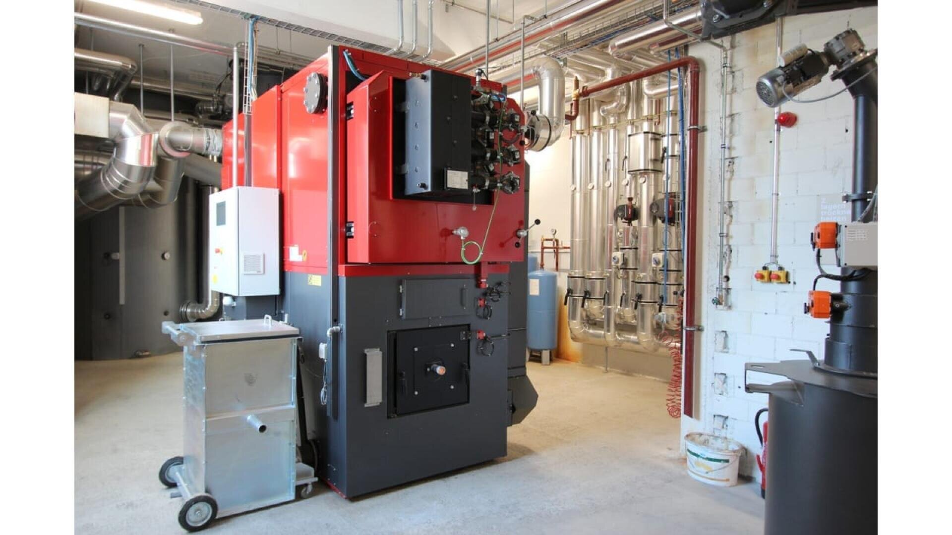 Une grande chaudière industrielle rouge et noire se trouve dans une salle de service propre avec des tuyaux apparents, des conduits métalliques, des panneaux de contrôle et un sol en béton.