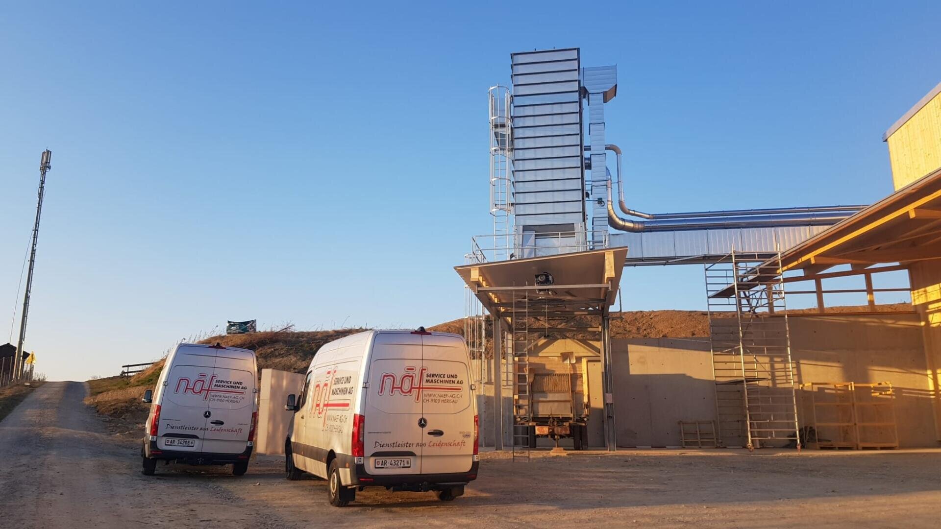 Zwei weiße Servicewagen parken auf einem unbefestigten Weg neben einer Industrieanlage mit großen metallenen Lüftungsschächten und einem hohen Gebäude, unter einem klaren blauen Himmel bei Sonnenuntergang.