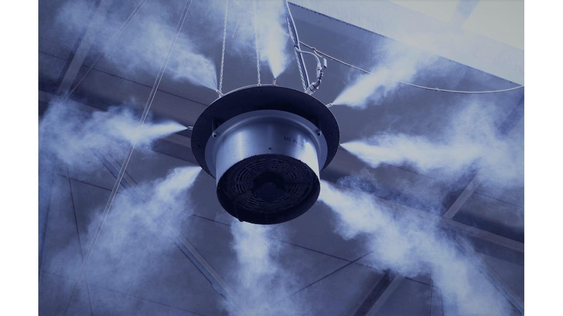 Un grand brumisateur circulaire suspendu au plafond pulvérise un brouillard fin ou de la vapeur dans de multiples directions, avec des poutres et des panneaux de toit visibles à l'arrière-plan.