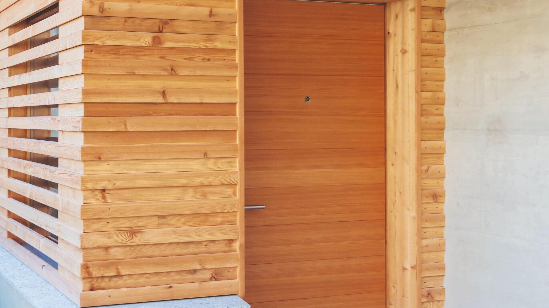 Eine moderne Holzeingangstür, die in eine Wand eingelassen ist, mit horizontalen Holzlatten auf der linken Seite und einer glatten Betonoberfläche auf der rechten Seite. Die Tür hat einen minimalistischen Griff und ein kleines Guckloch.
