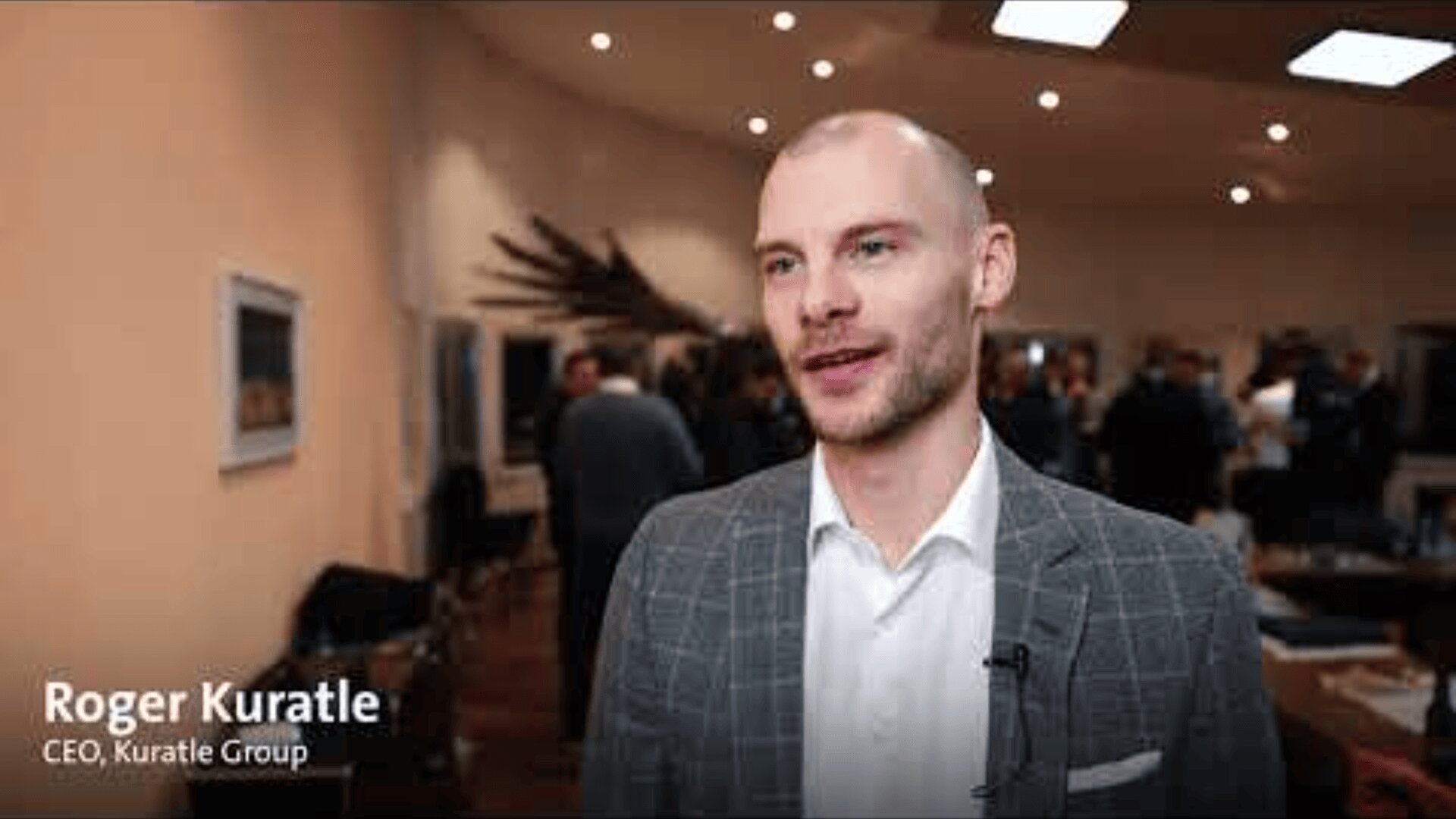 Ein Mann in einem grau karierten Anzug steht bei einer Veranstaltung in einem Innenraum, spricht und lächelt. Der Text auf dem Bild lautet: Roger Kuratle, CEO, Kuratle Gruppe. Im unscharfen Hintergrund sind Menschen und Kunstwerke zu sehen.