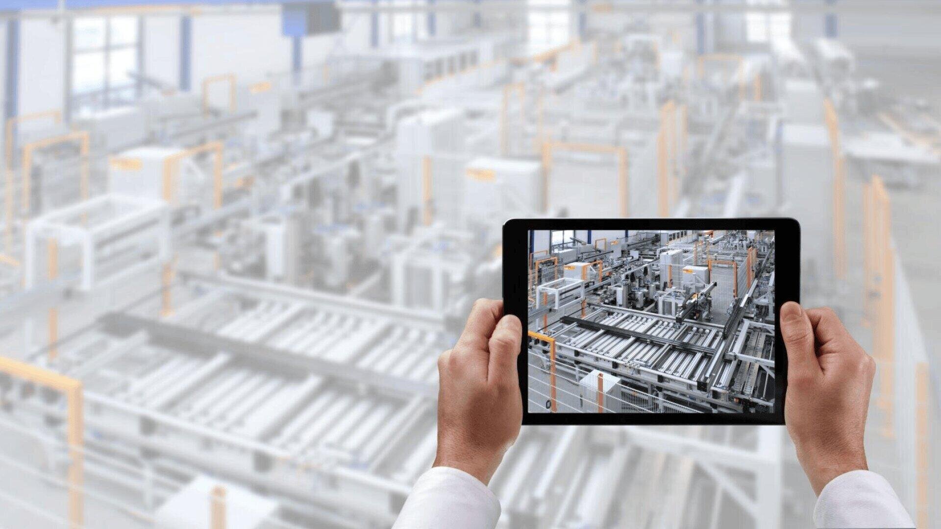 Une personne tient une tablette montrant une image claire et détaillée d'une usine automatisée ou d'une chaîne de production industrielle, tandis que l'arrière-plan montre le même sol d'usine flou.