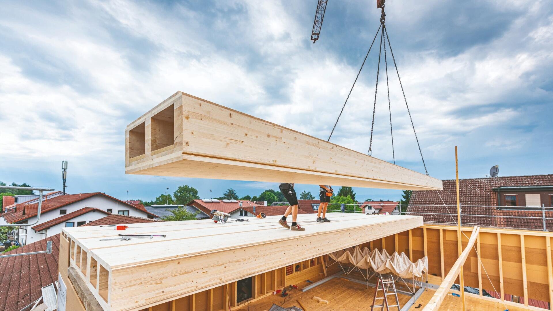 Ein Kran hebt einen großen Holzbalken in das obere Stockwerk eines im Bau befindlichen Gebäudes. Arbeiter stehen auf dem Dach und führen den Balken in die richtige Position. Im Hintergrund sind Wohnhäuser und der bewölkte Himmel zu sehen.