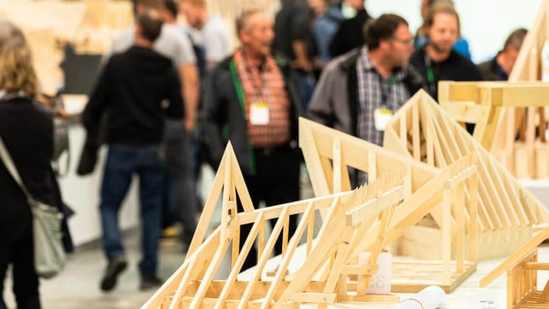 Auf einem Tisch im Vordergrund sind hölzerne Architekturmodelle ausgestellt, während sich im unscharfen Hintergrund eine Gruppe von Menschen in Freizeitkleidung unterhält und umhergeht, was auf eine Ausstellung oder Veranstaltung hindeutet.