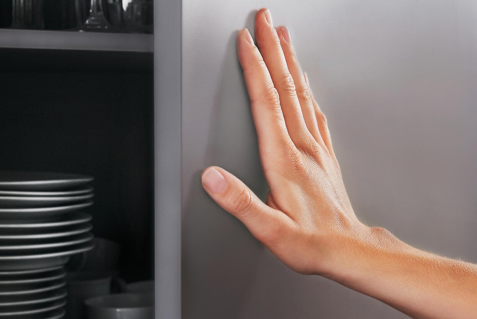 Une main touche l'extérieur lisse d'une porte d'armoire fermée, à côté d'une étagère ouverte sur laquelle sont empilés des assiettes et des bols.