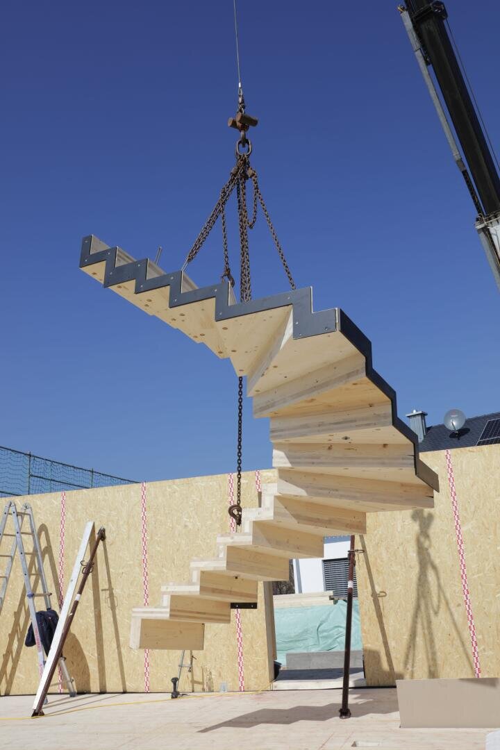 Eine große Holztreppe wird von einem Kran auf einer Baustelle hochgezogen. Die Treppe ist mit Ketten in der Luft aufgehängt, und im Hintergrund sind unfertige Holzwände und Bauwerkzeuge zu sehen.