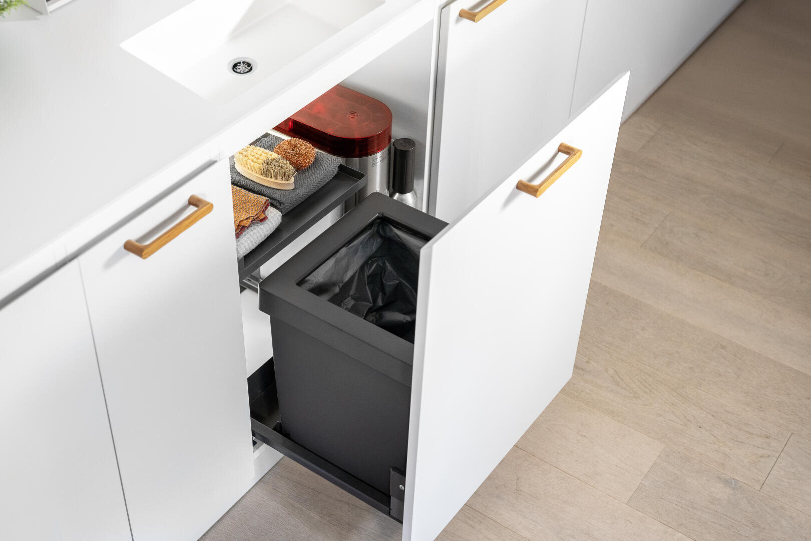 Ein offener weißer Küchenschrank zeigt einen ausziehbaren Mülleimer mit schwarzem 40-Liter-Behälter und darüber ein fixes Regal mit Reinigungsschwämmen, einer Bürste. Daneben ein Wasseraufbereitungsgerät. Der Schrank steht unter einer weißen Arbeitsplatte mit einem Waschbecken.