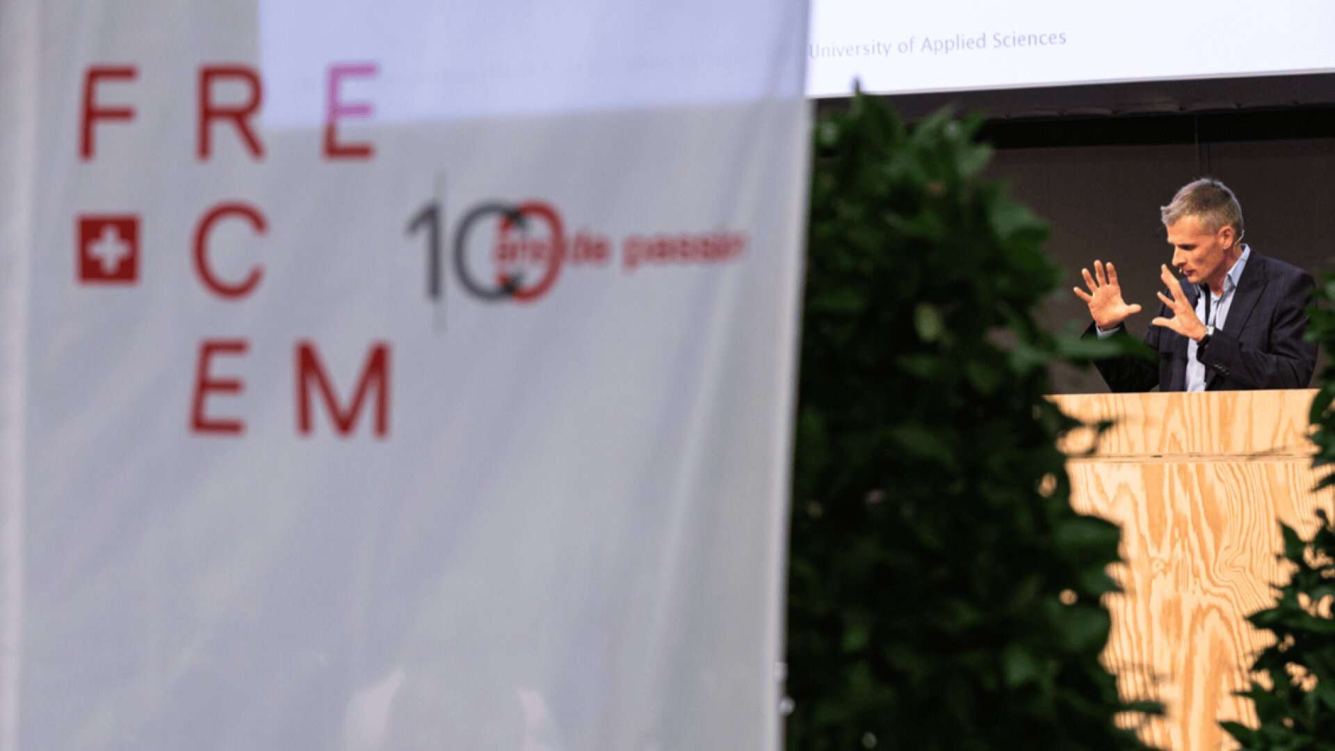 Ein Mann steht an einem hölzernen Podium, spricht und gestikuliert mit seinen Händen. Im Vordergrund verdecken ein verschwommenes Banner mit roten Buchstaben, auf dem FRECEM steht, und Grünpflanzen teilweise die Sicht.