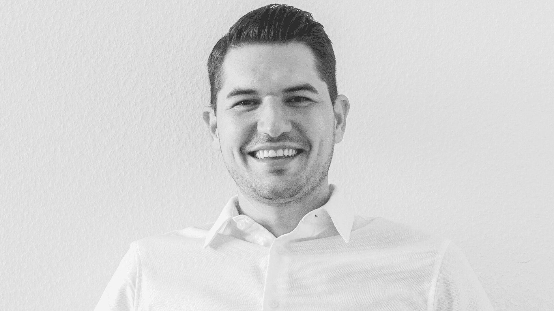 Ein Mann mit kurzen dunklen Haaren und einem hellen Hemd sitzt lächelnd vor einem schlichten hellen Hintergrund. Das Bild ist schwarz-weiß.