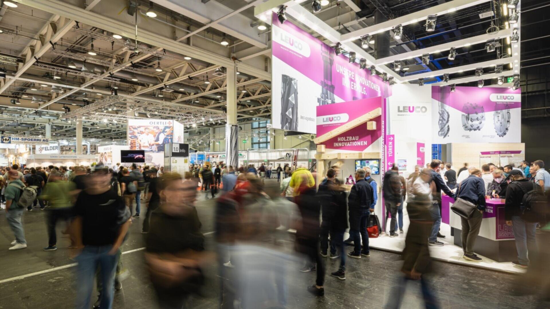 Un salon professionnel très fréquenté, avec de nombreuses personnes marchant et se tenant debout près de stands très éclairés, dont un stand LEUCO rose et blanc bien en vue. Le hall est spacieux, avec de hauts plafonds et des poutres apparentes.