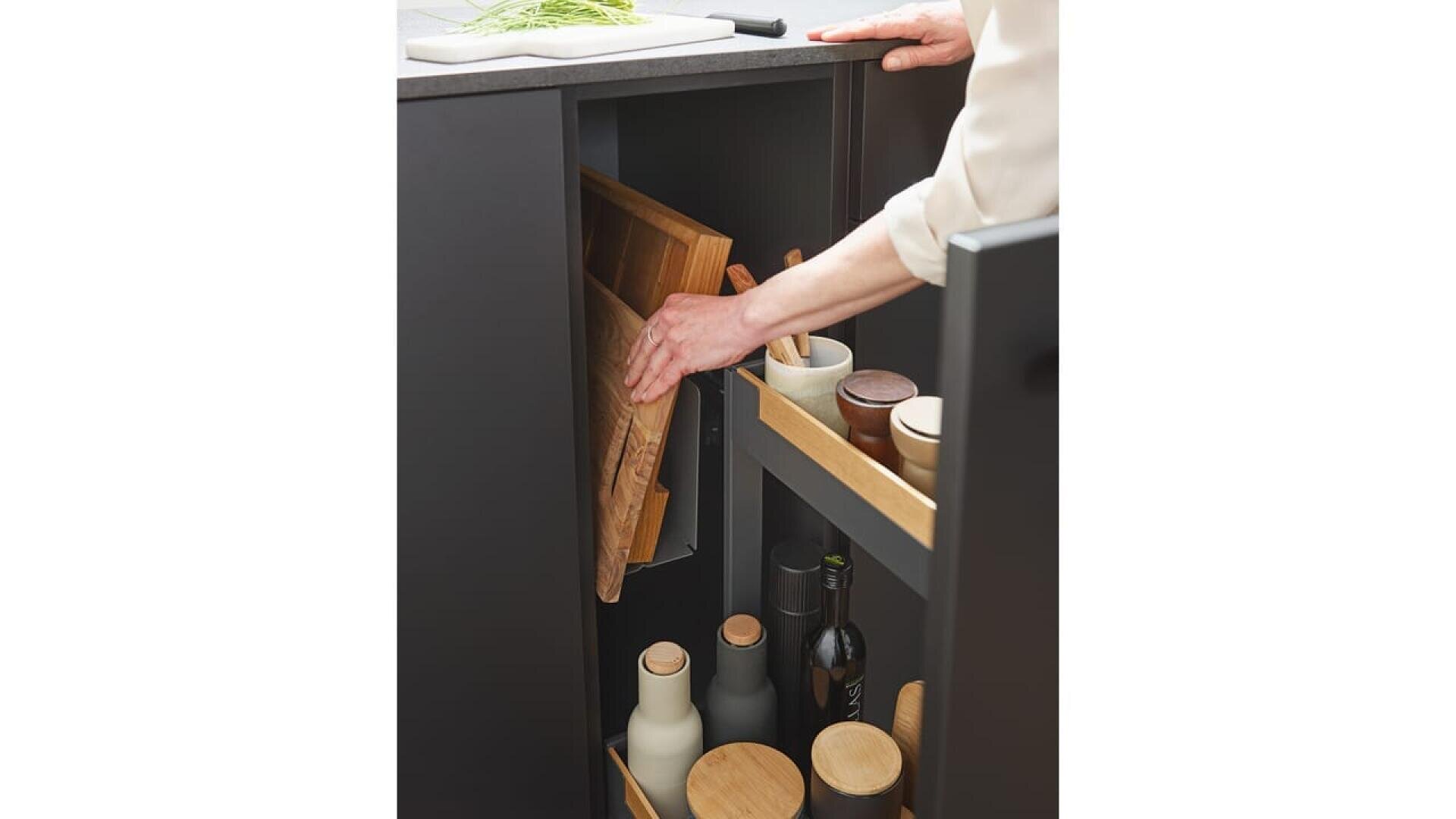 Une personne place une planche à découper en bois dans une fente verticale à l'intérieur d'un meuble de cuisine noir, à côté d'étagères contenant des bouteilles, des bocaux et des récipients.