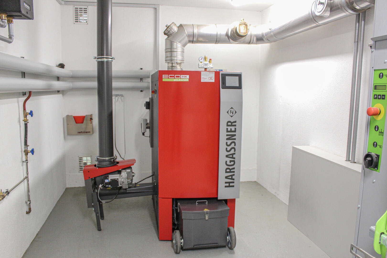 In einem sauberen, weißen Hauswirtschaftsraum ist eine rote Hargassner ECO-Heizkesseleinheit mit Metallrohren und einem Bedienfeld an der Wand daneben installiert. Unter dem Kessel befindet sich ein schwarzer Aschebehälter.