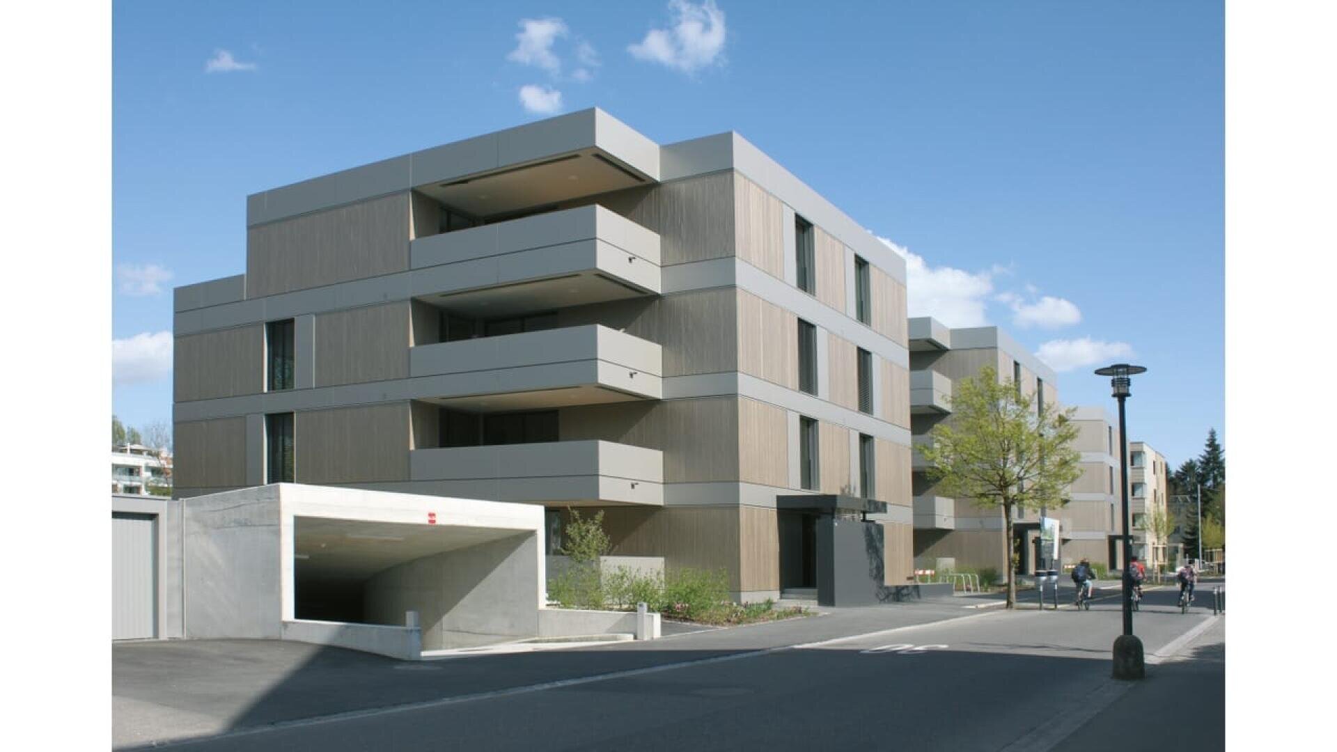 Immeuble d'appartements moderne au design minimaliste, avec de grands balcons et des toits plats, situé dans une rue propre avec des arbres et un ciel bleu au-dessus de la tête. Un parking couvert est visible au premier plan.