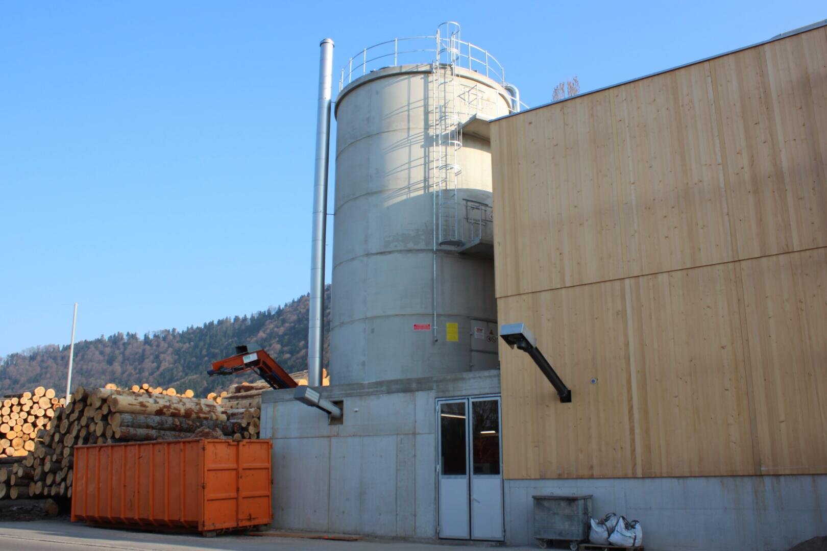 Ein großer zylindrischer Betonbau mit Metalltreppen steht neben einem Holzgebäude; an einem sonnigen Tag sind draußen orangefarbene Müllcontainer und gestapelte Holzscheite zu sehen.