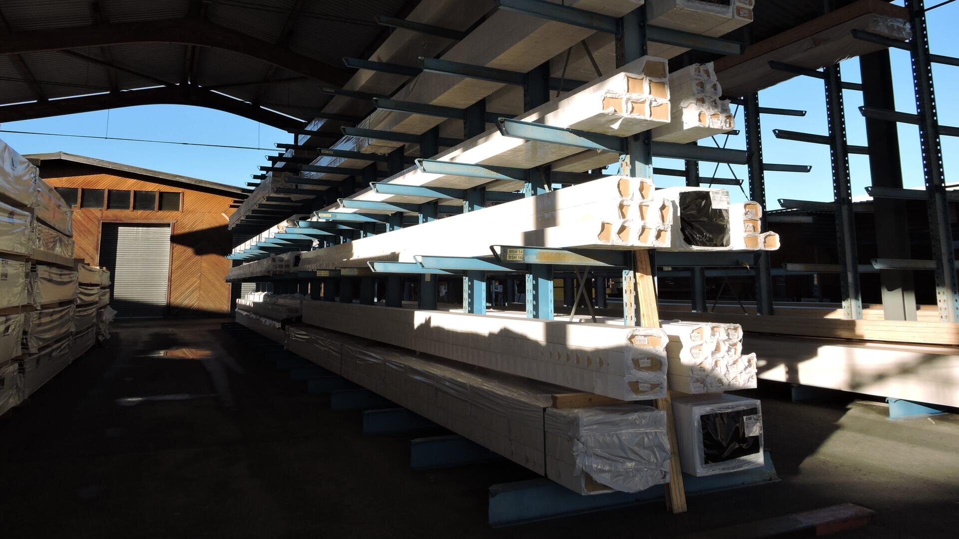 In einer offenen Lagerhalle, in der das Sonnenlicht Schatten auf den Boden wirft, lagern auf Metallregalen Stapel von langen, mit Plastik umwickelten Holzbalken.