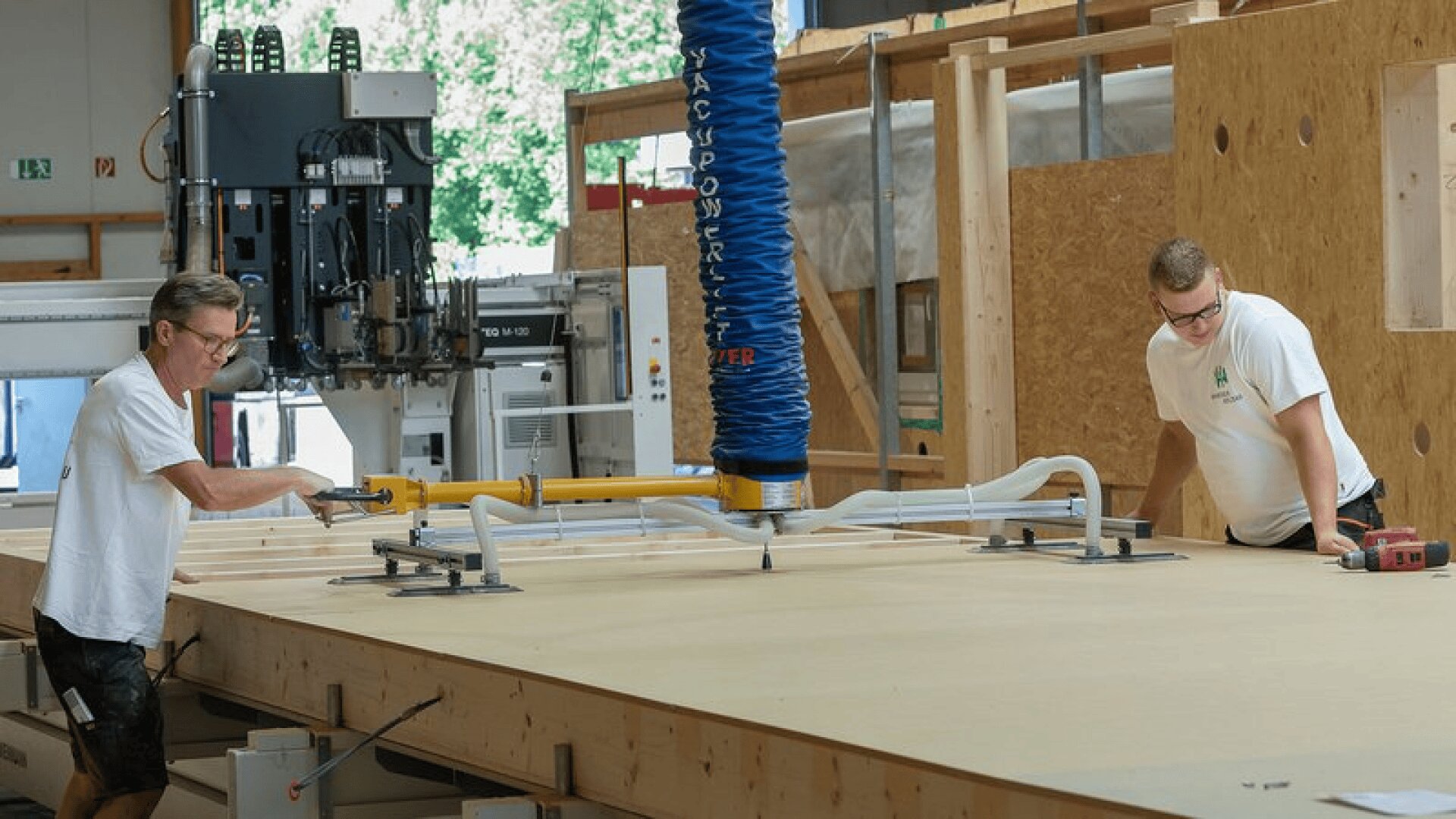 Zwei Männer in einer Werkstatt heben mit einer großen Saugmaschine eine Holzplatte auf einen Tisch. Beide tragen weiße Hemden und Brillen, und in dem Raum befinden sich Holzplatten und industrielle Geräte.