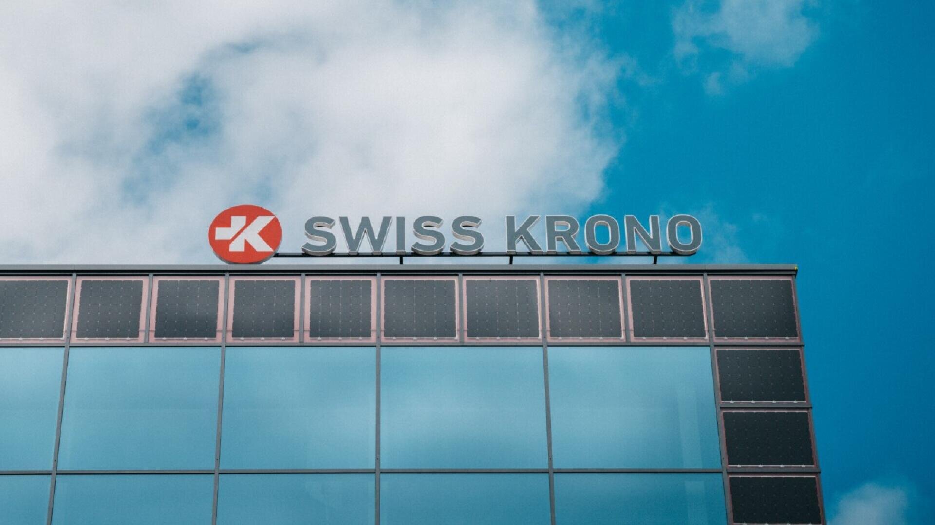 Un bâtiment moderne en verre surmonté du logo et du texte SWISS KRONO, dans un ciel bleu partiellement nuageux.