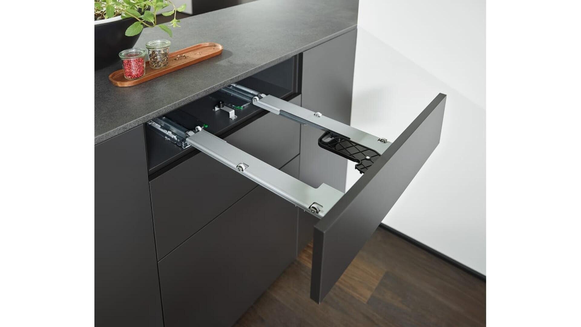 Un meuble de cuisine moderne avec un mécanisme de tiroir coulissant déployé, montrant les glissières et la quincaillerie en métal. Deux pots à épices et une plante sont posés sur le plan de travail.