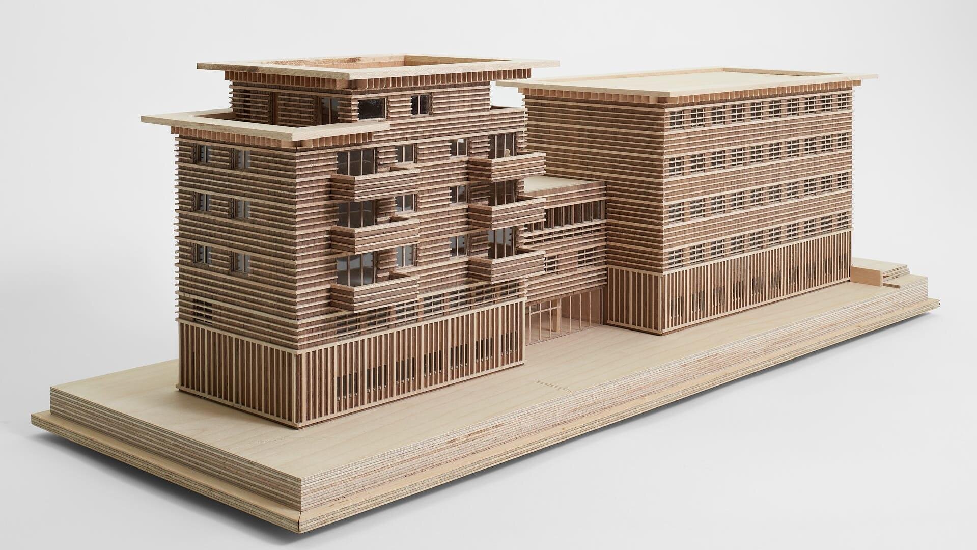 Modèle architectural détaillé en bois d'un bâtiment moderne à plusieurs étages avec des lattes horizontales et des formes rectangulaires, présenté sur une base plate en bois sur un fond blanc uni.