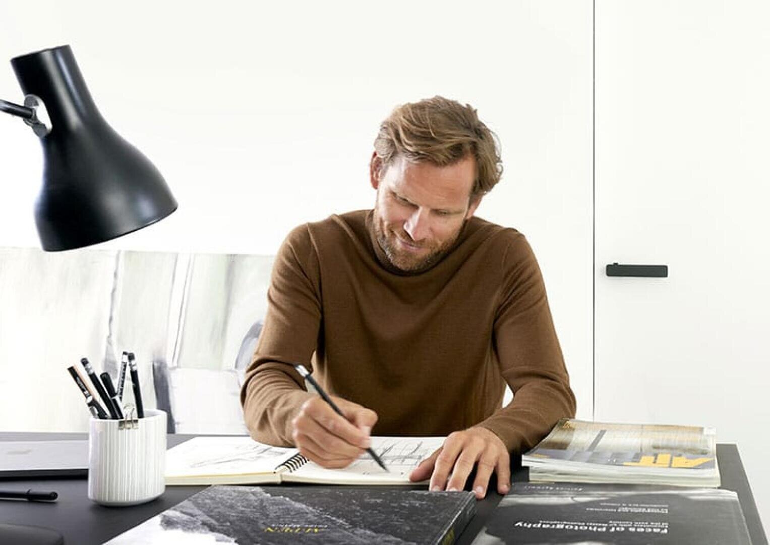 Ein Mann mit hellbraunem Haar und Bart, der einen braunen Pullover trägt, sitzt an einem Schreibtisch und zeichnet in ein Notizbuch. Auf dem Schreibtisch liegen Bücher, Stifte und eine schwarze Lampe. Der Hintergrund ist minimal und hell.