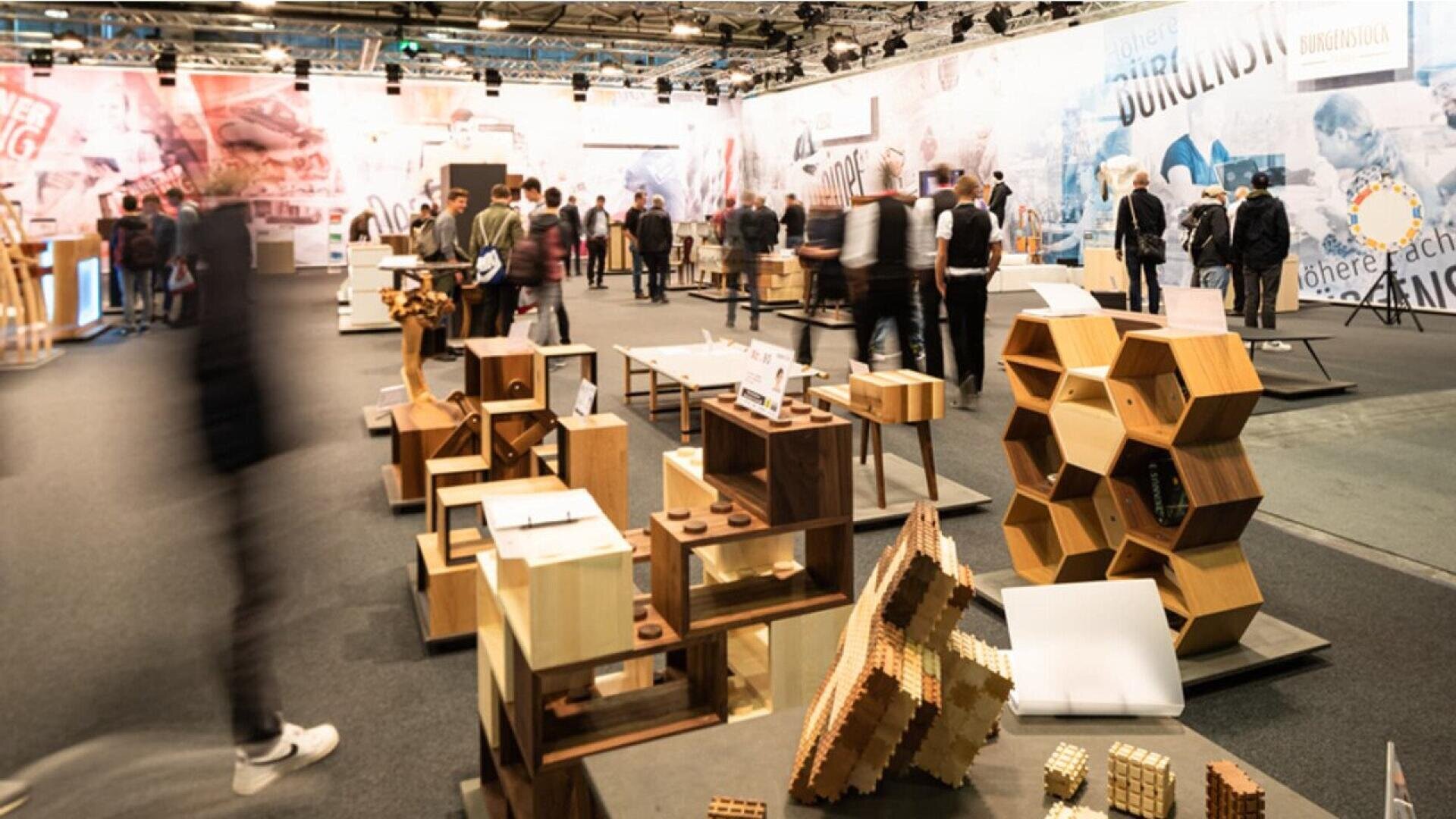 Un hall d'exposition très fréquenté, avec des gens qui se promènent et regardent des expositions de meubles et de sculptures modernes et géométriques en bois, disposés sur des stands, avec en toile de fond de grands graphiques muraux.