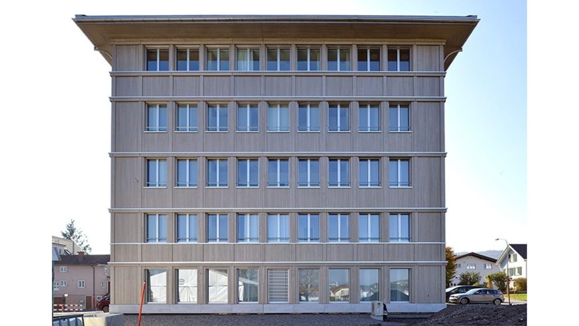 Un bâtiment moderne de six étages avec un toit plat et une façade en bois clair, avec des rangées de fenêtres rectangulaires identiques. En arrière-plan, des petites maisons et un ciel dégagé.